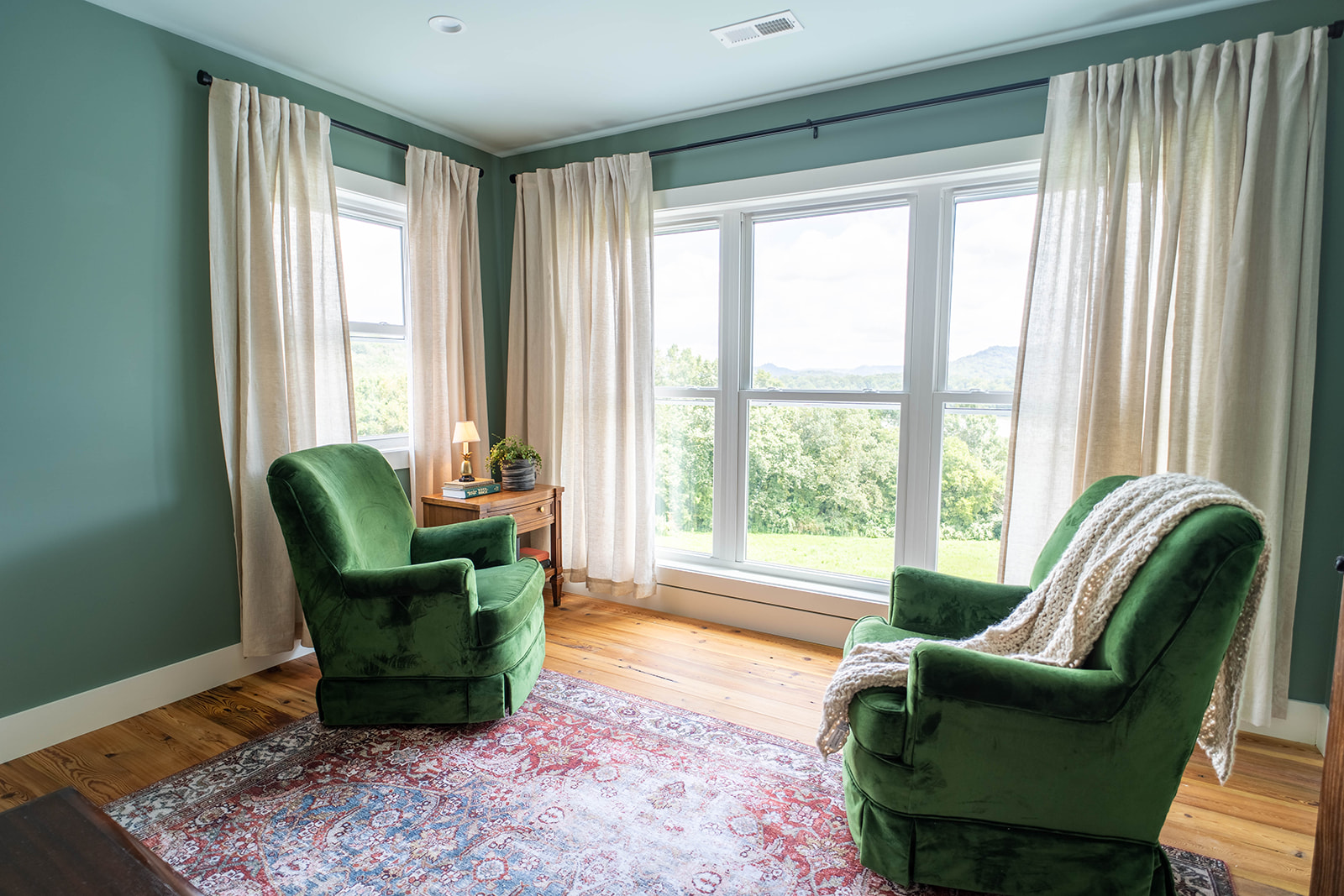 Cozy reading nook featuring two green velvet armchairs, a wooden side table with a lamp, and large windows with sheer curtains, overlooking a lush landscape. The room has a calming green wall and a patterned area rug on wooden flooring.