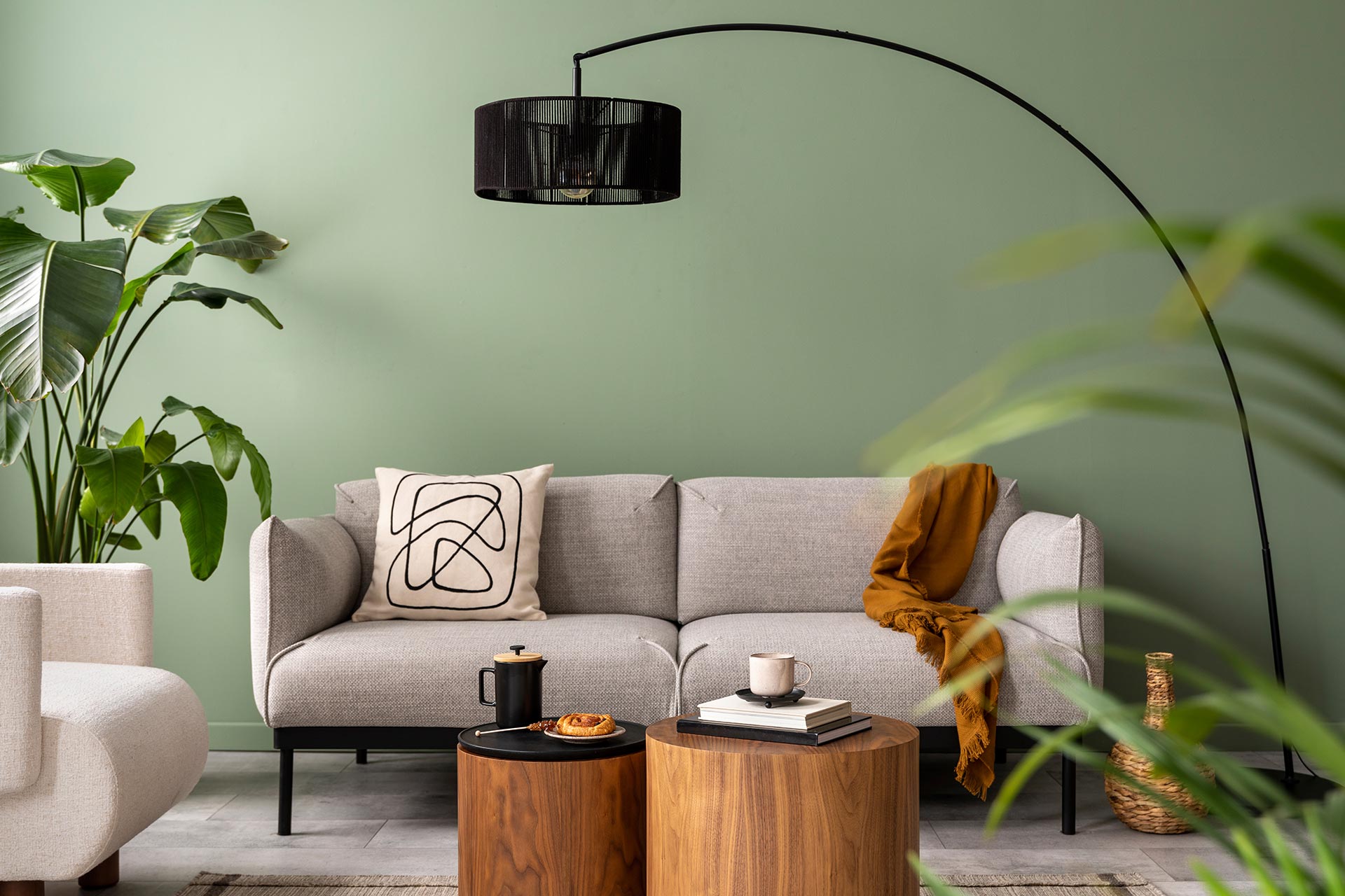 Modern living room interior featuring a gray sofa with an abstract patterned pillow, a stylish black arc floor lamp, and wooden side tables. The space is accented with green walls and lush indoor plants, creating a cozy and inviting atmosphere.